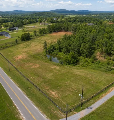 Aerial view of a rural land parcel showing road access, utility lines, nearby homes, trees, terrain, and a pond—key factors that can affect land value.